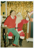 Man wearing a red sweater and green slacks sitting smiling on Santa's lap with festive gold decorations in the background.